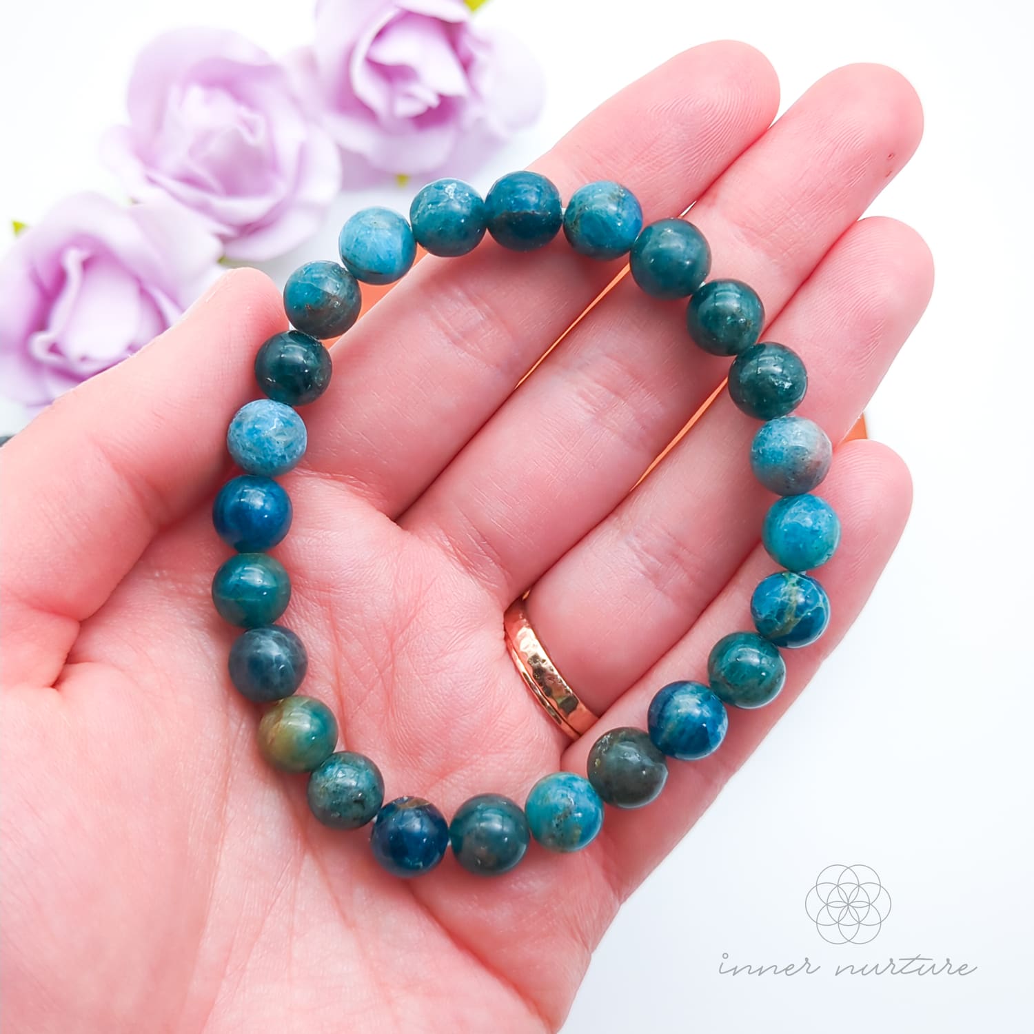 Blue beaded bracelet on a wooden disc with pink flowers in the background. Blue apatite 8mm bracelet sold by Inner Nurture, an online crystal shop in Australia.