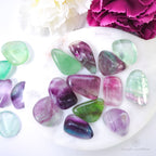 rainbow fluorite tumbles on white plate with pink and white flowers in the background. available online at inner nurture, an online crystal shop in australia