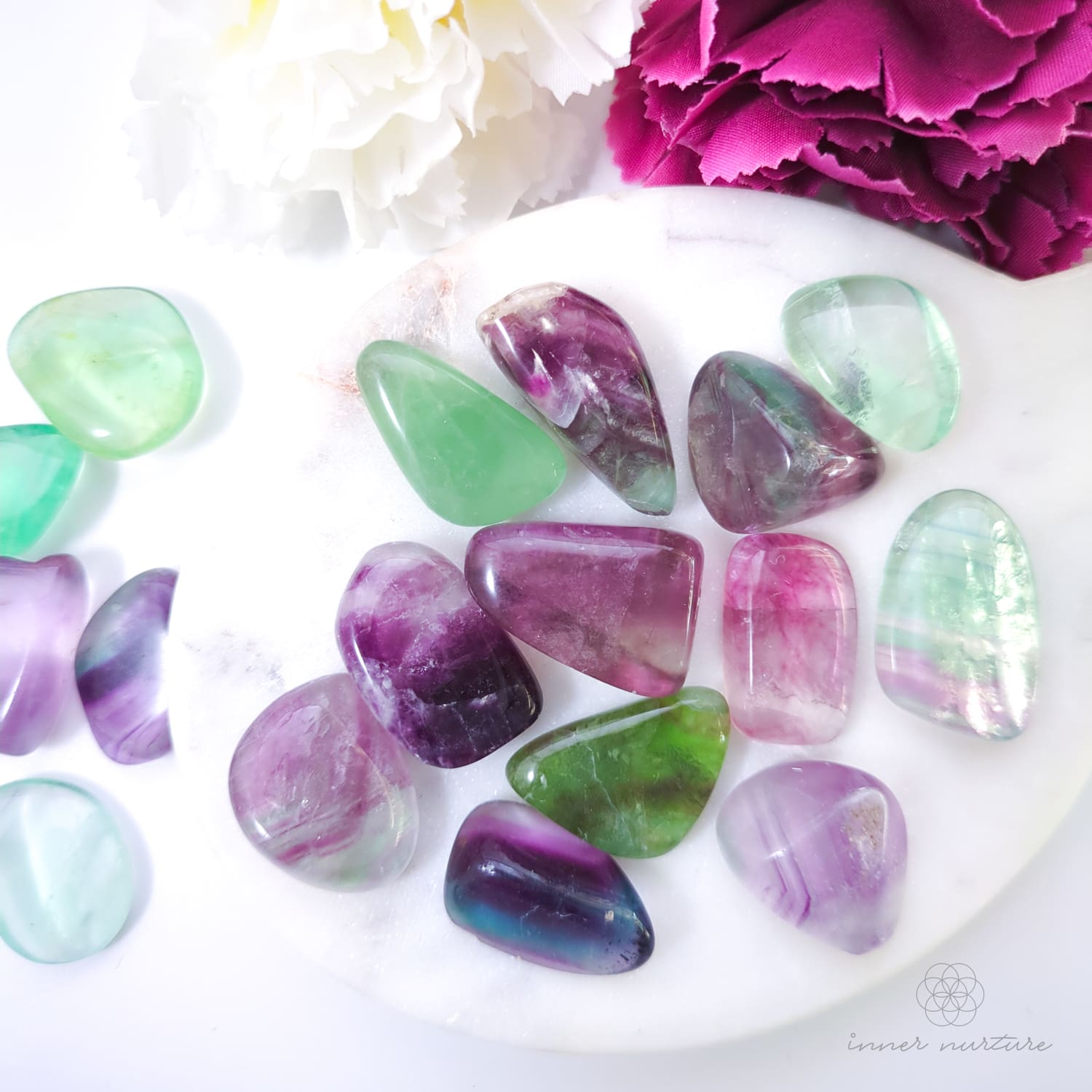 rainbow fluorite tumbles on white plate with pink and white flowers in the background. available online at inner nurture, an online crystal shop in australia