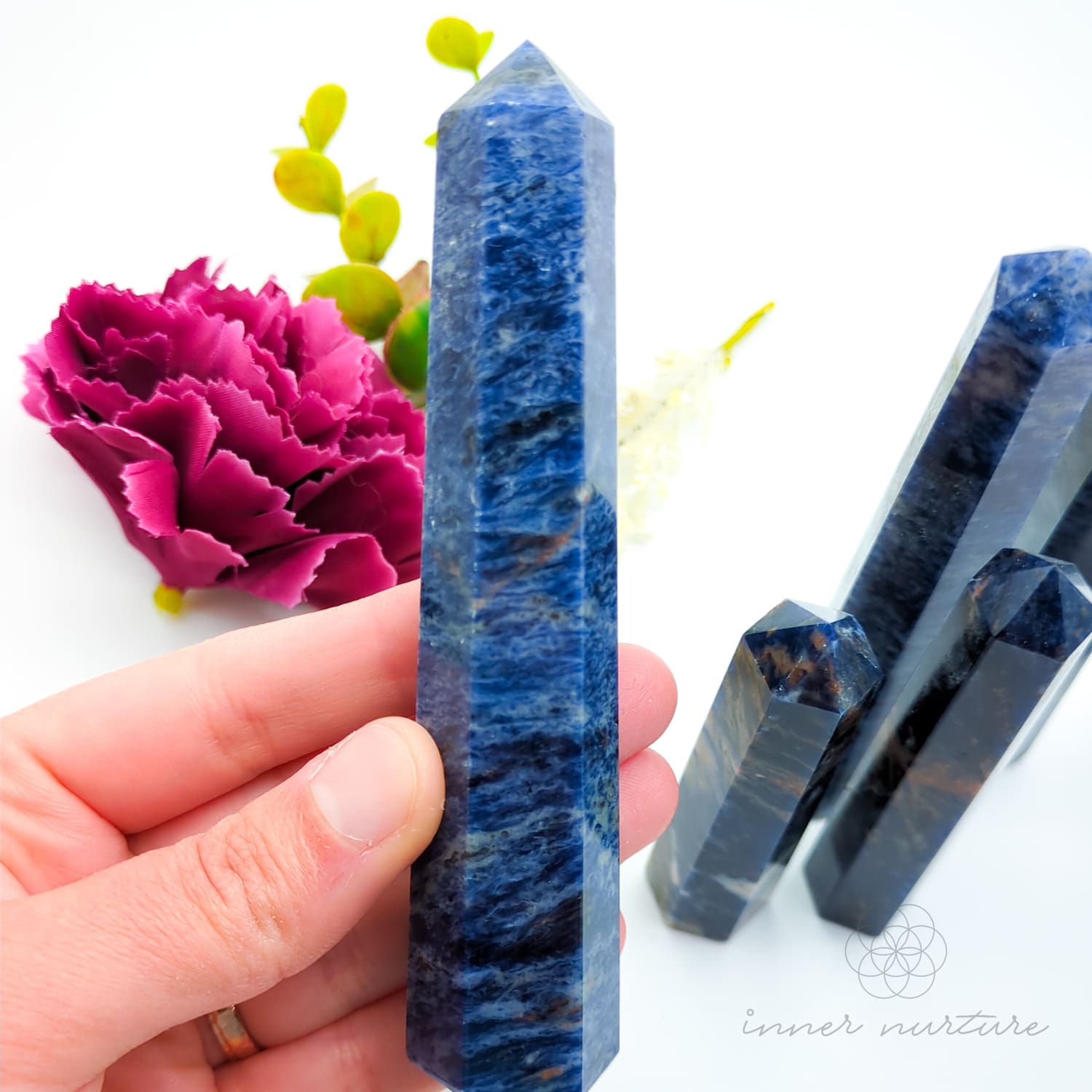 Blue crystal held by a hand with pink flower and green leaves in the background. Sodalite Generator Tower available to Buy from Inner Nurture - an online Crystals shop in Australia