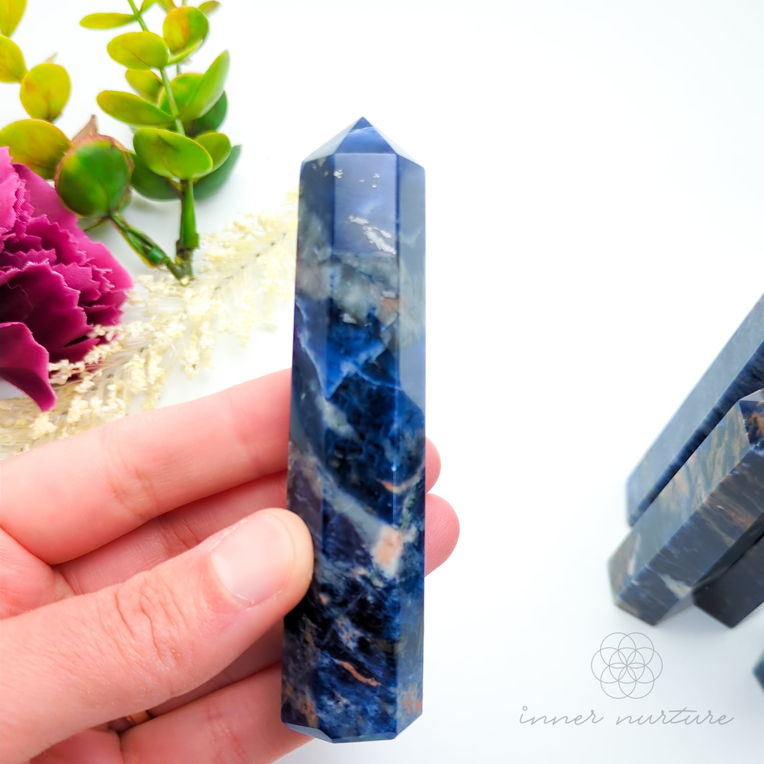 Blue crystal tower held in a hand with a blurred background of flowers and stones. Sodalite Generator Tower available to Buy from Inner Nurture - an online Crystals shop in Australia