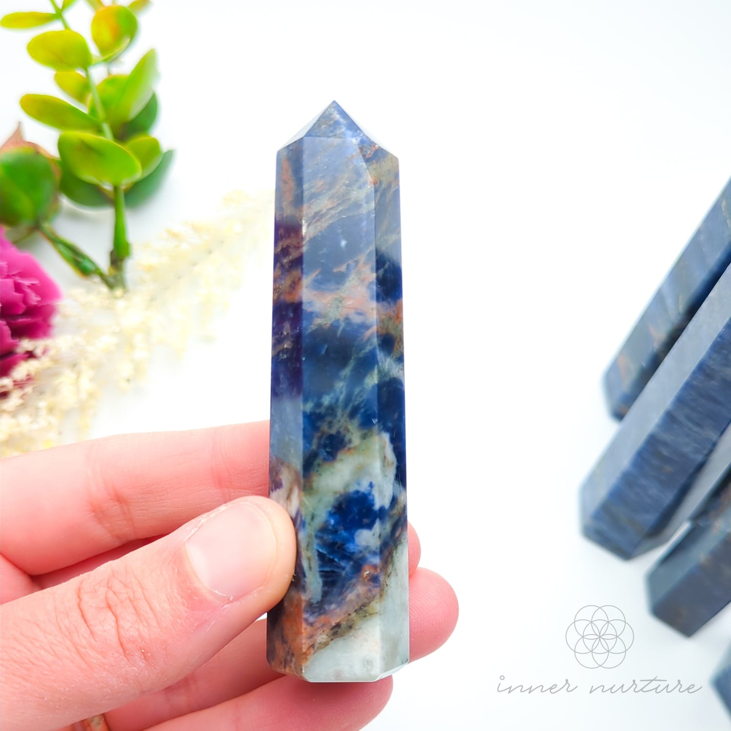 Blue crystal tower held in a hand with a blurred background of flowers and stones. Sodalite Generator Tower available to Buy from Inner Nurture - an online Crystals shop in Australia