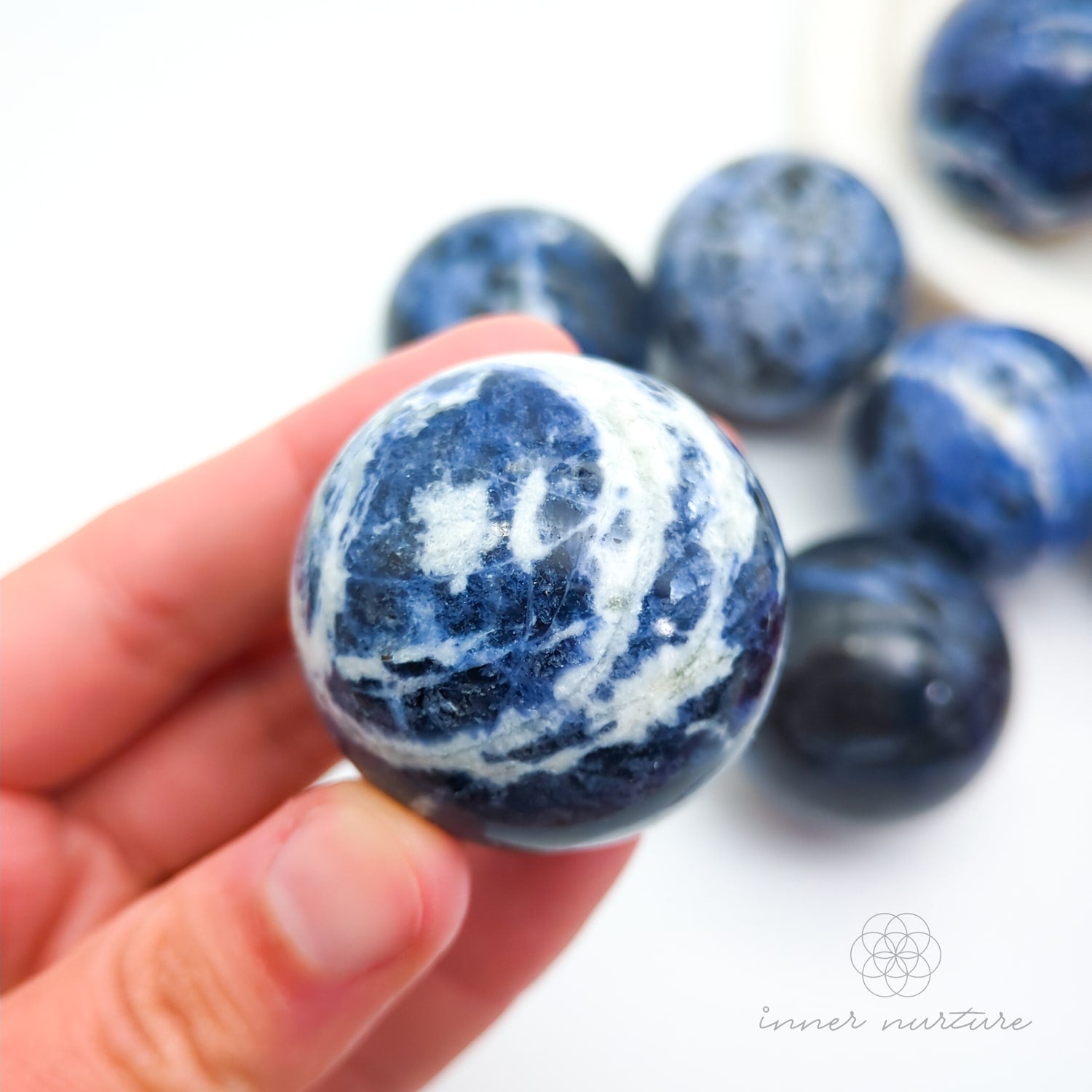 Blue and white marbled stones held in a hand with more stones in the background. Sodalite Sphere sold at inner nurture, an australian online crystal shop