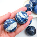 Blue and white marbled stones held in a hand with more stones in the background. Sodalite Sphere sold at inner nurture, an australian online crystal shop