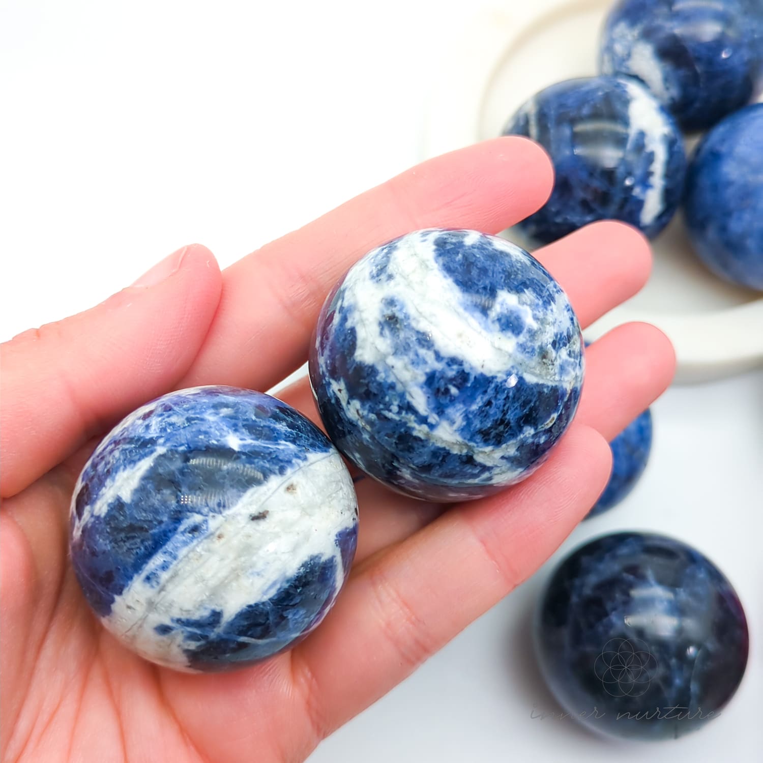Blue and white marbled stones held in a hand with more stones in the background. Sodalite Sphere sold at inner nurture, an australian online crystal shop