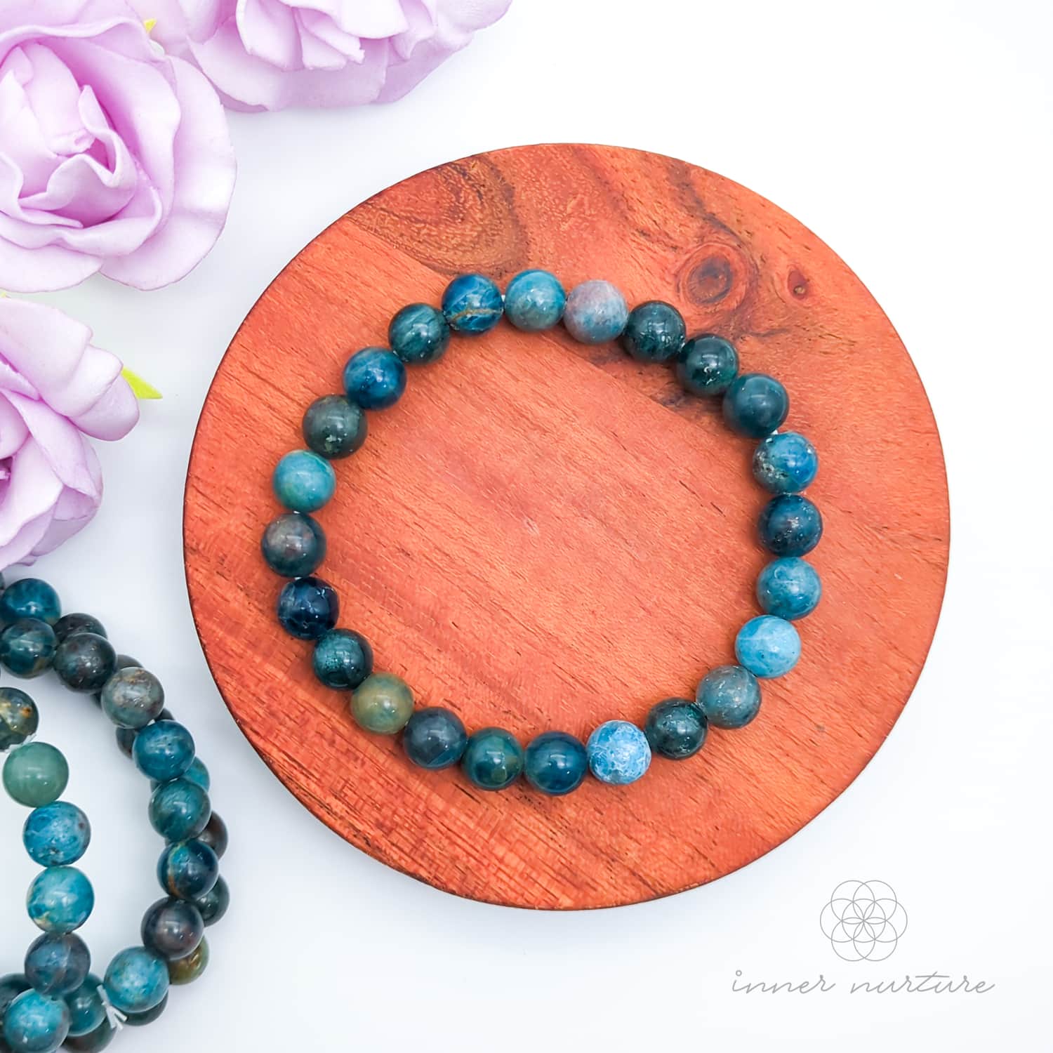 Blue beaded bracelet on a wooden disc with pink flowers in the background. Blue apatite 8mm bracelet sold by Inner Nurture, an online crystal shop in Australia. 