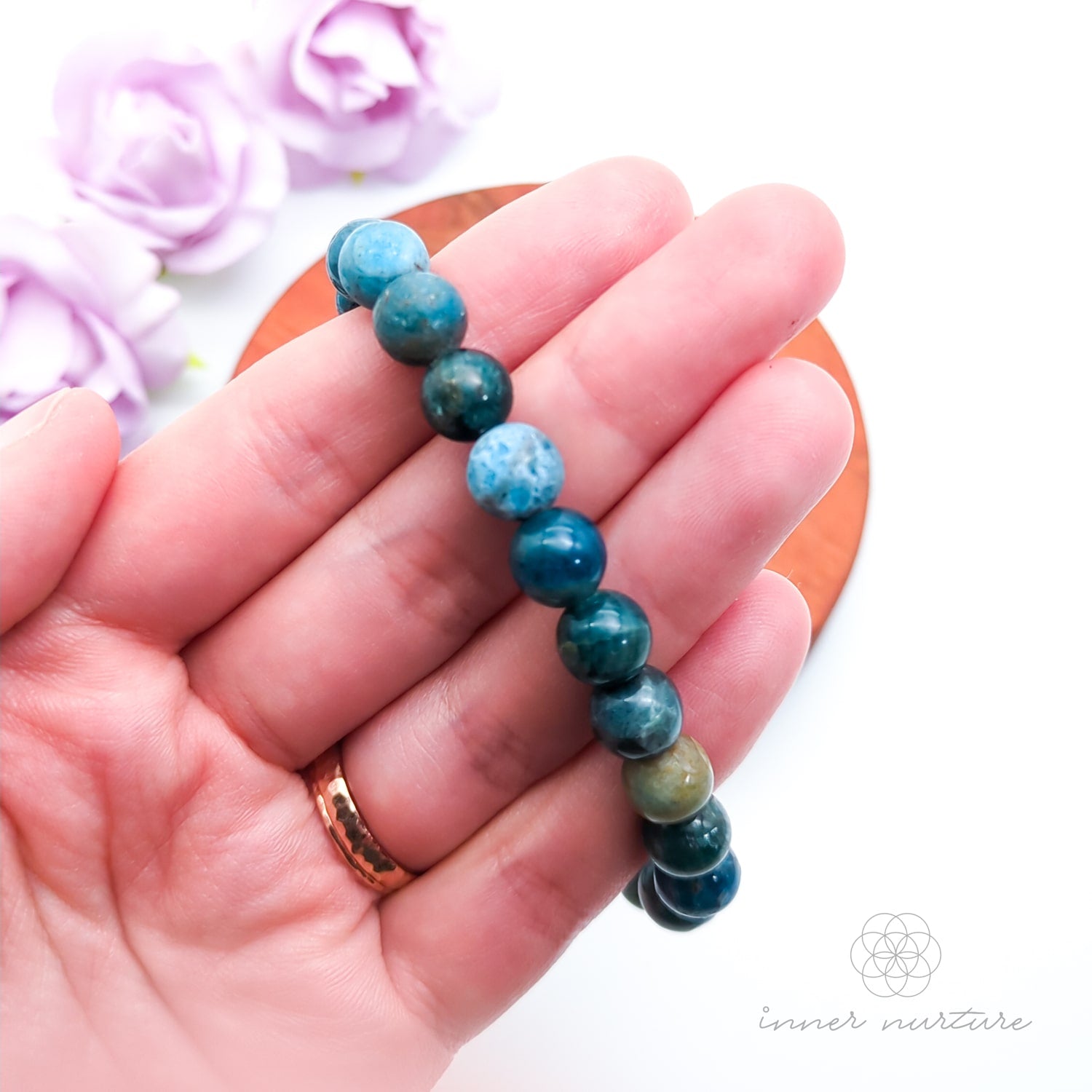 Blue beaded bracelet on a wooden disc with pink flowers in the background. Blue apatite 8mm bracelet sold by Inner Nurture, an online crystal shop in Australia. 