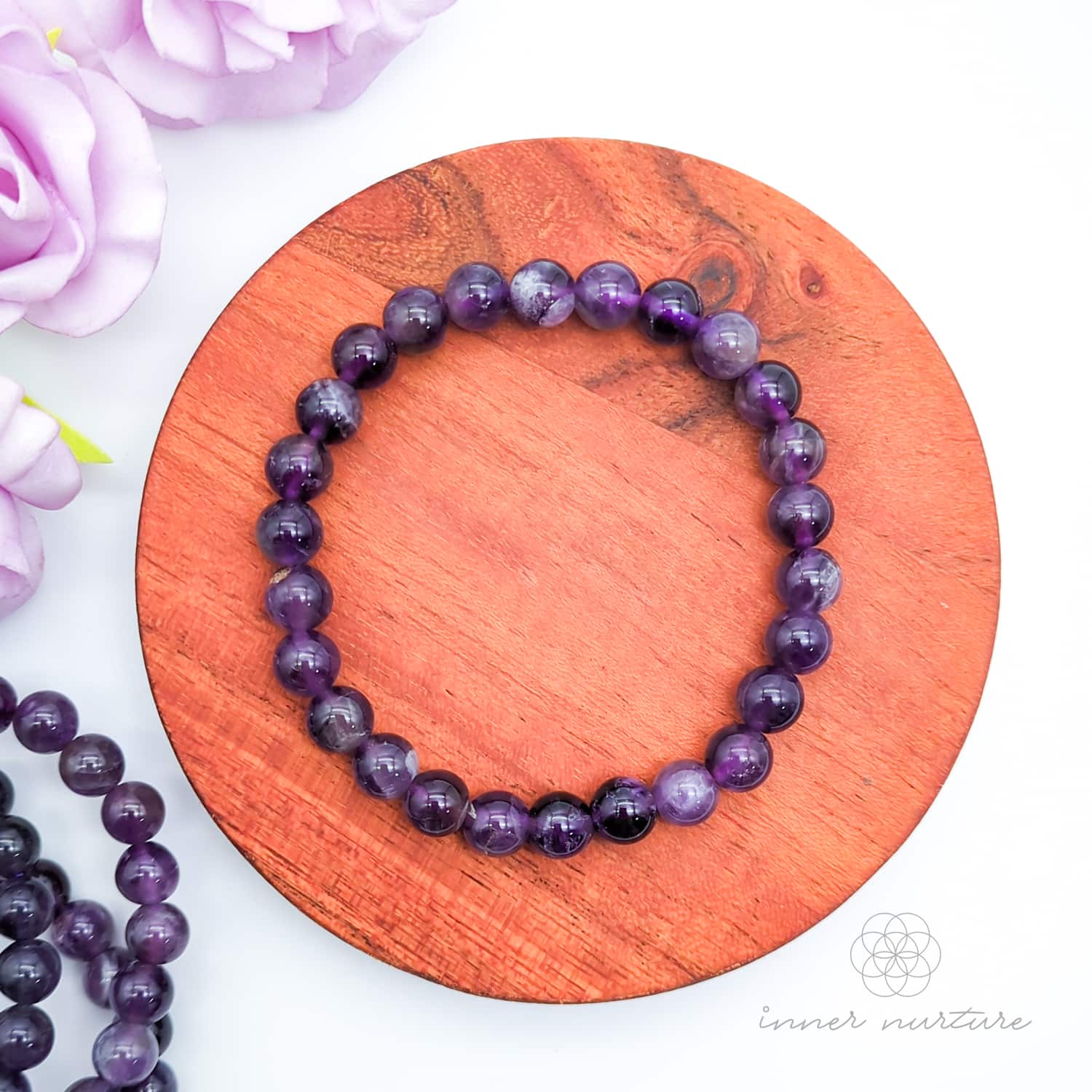 Purple beaded bracelet on a wooden surface with flowers in the background. Multicolored beaded bracelet on a wooden surface with flowers in the background. Blue beaded bracelet on a wooden disc with pink flowers in the background. Amethyst bead bracelet sold by Inner Nurture, an online crystal shop in Australia. 
