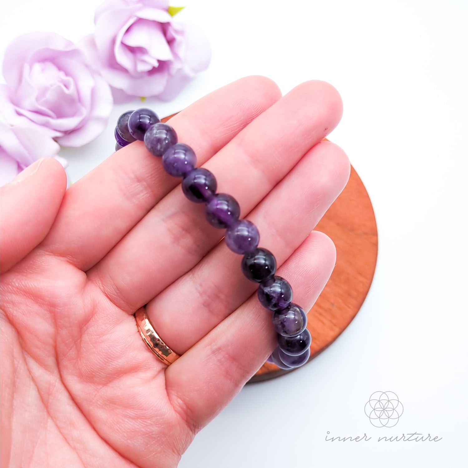 Purple beaded bracelet on a wooden surface with flowers in the background. Multicolored beaded bracelet on a wooden surface with flowers in the background. Blue beaded bracelet on a wooden disc with pink flowers in the background. Amethyst bead bracelet sold by Inner Nurture, an online crystal shop in Australia. 