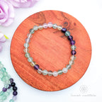 Multicolored beaded bracelet on a wooden surface with flowers in the background. Blue beaded bracelet on a wooden disc with pink flowers in the background. Rainbow Fluorite bead bracelet sold by Inner Nurture, an online crystal shop in Australia. 