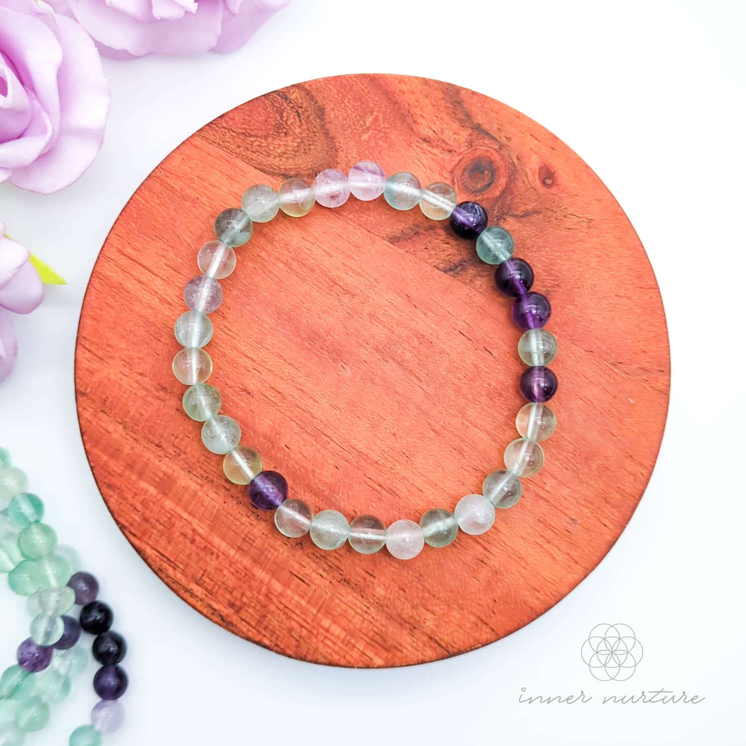 Multicolored beaded bracelet on a wooden surface with flowers in the background. Blue beaded bracelet on a wooden disc with pink flowers in the background. Rainbow Fluorite bead bracelet sold by Inner Nurture, an online crystal shop in Australia. 