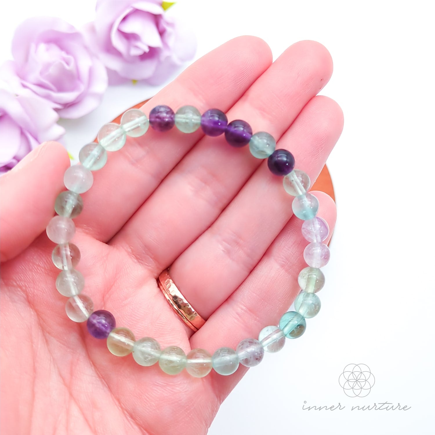 Multicolored beaded bracelet on a wooden surface with flowers in the background. Blue beaded bracelet on a wooden disc with pink flowers in the background. Rainbow Fluorite bead bracelet sold by Inner Nurture, an online crystal shop in Australia. 