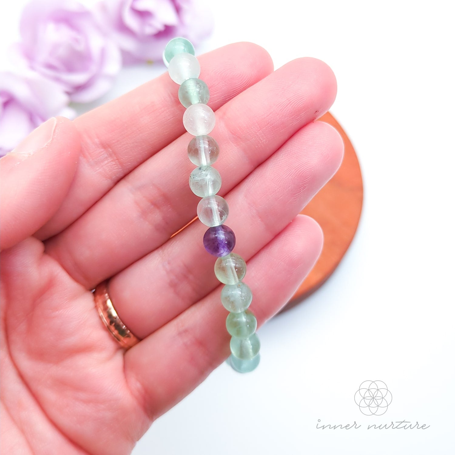 Multicolored beaded bracelet on a wooden surface with flowers in the background. Blue beaded bracelet on a wooden disc with pink flowers in the background. Rainbow Fluorite bead bracelet sold by Inner Nurture, an online crystal shop in Australia. 