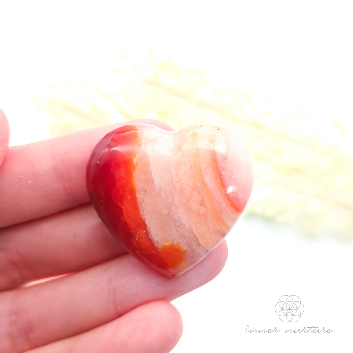 Carnelian crystal heart held in a hand with yellow flowers in the backdrop. Sold at Inner Nurture, an online crystal shop in Australia. 