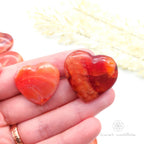 Carnelian crystal hearts held in a hand with yellow flowers in the backdrop. Sold at Inner Nurture, an online crystal shop in Australia. 