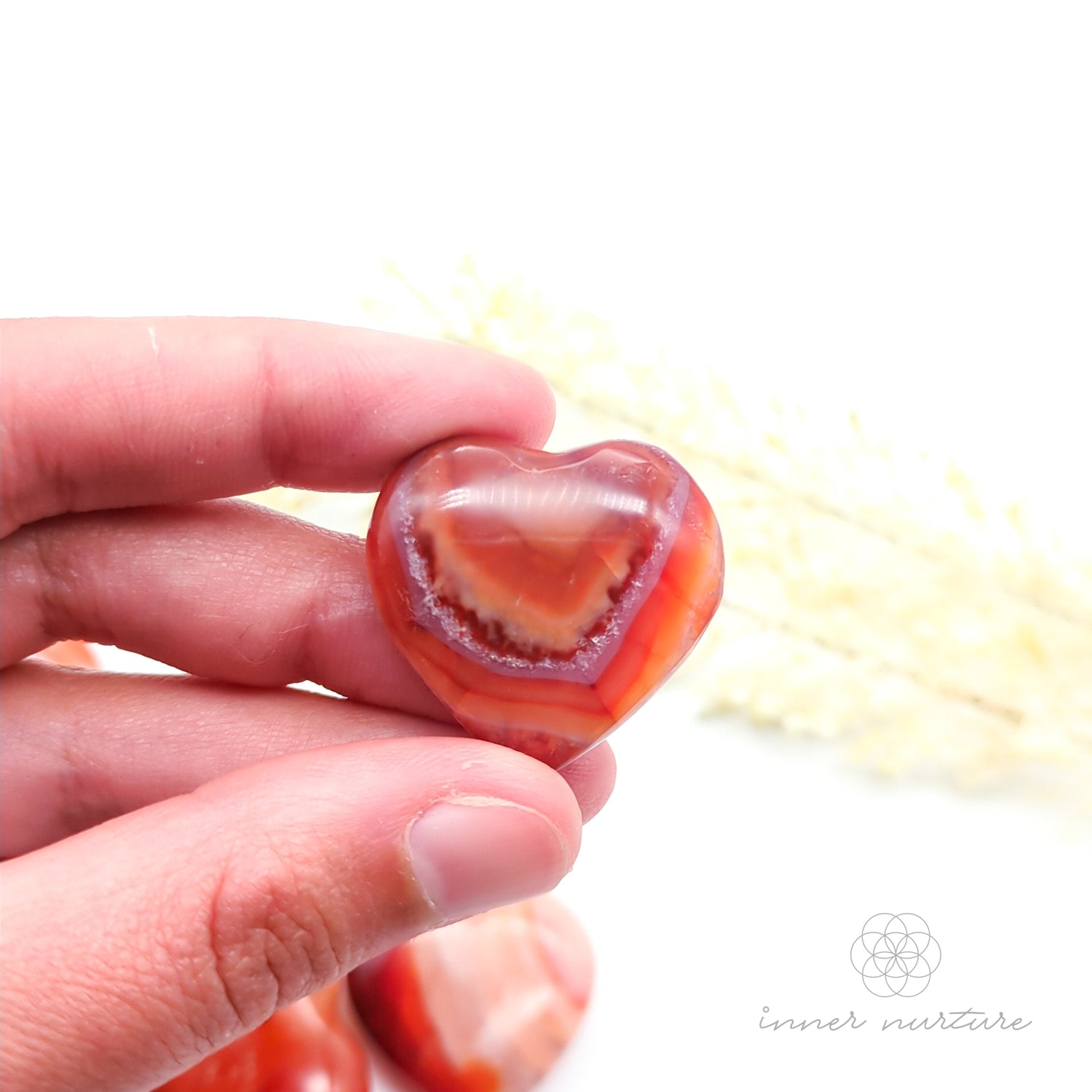 Carnelian crystal heart held in a hand with yellow flowers in the backdrop. Sold at Inner Nurture, an online crystal shop in Australia. 