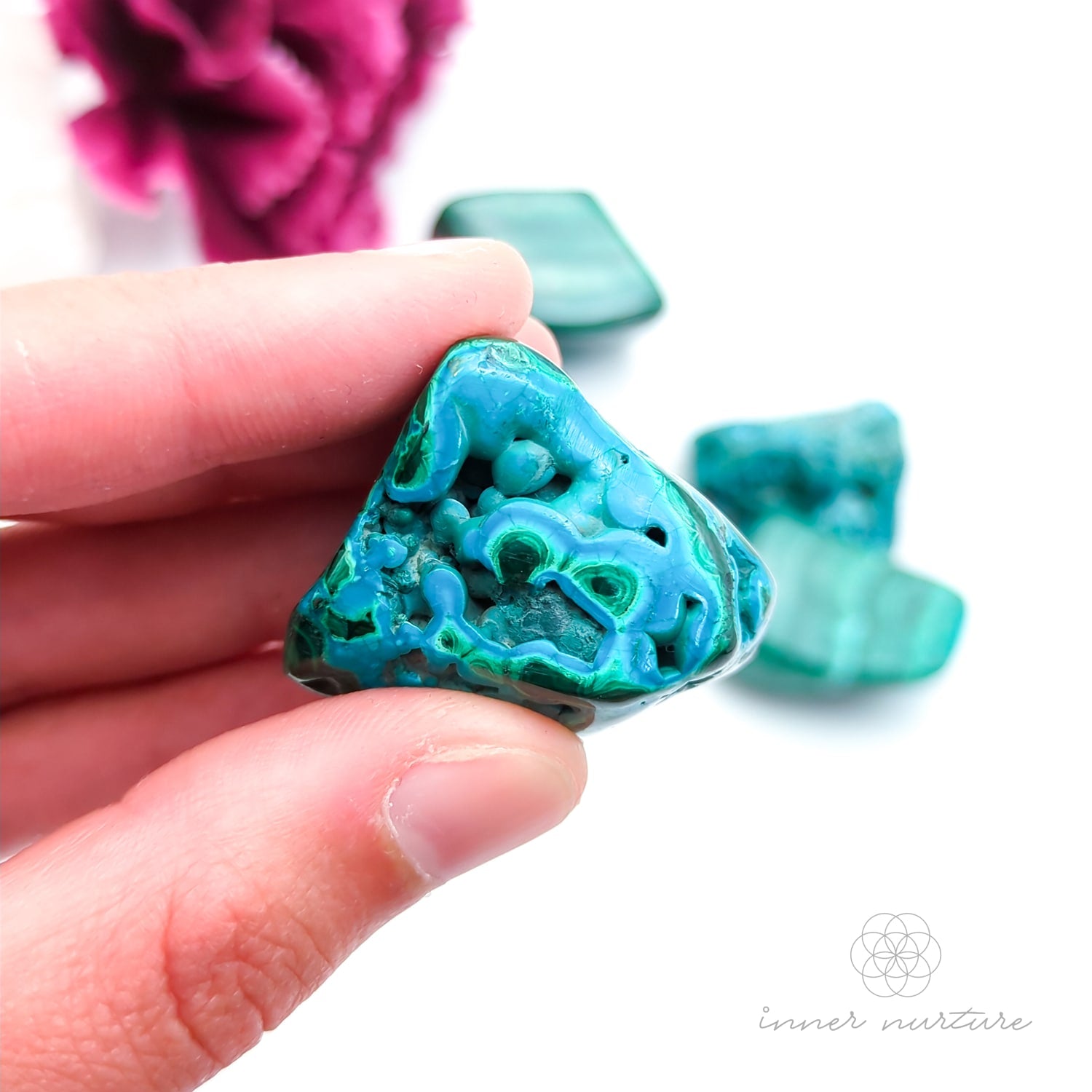 Blue green stone held in hand, with more stones in the background and a white and purple flower. Malacolla sold at Inner Nurture, an online crystal shop in Australia. 