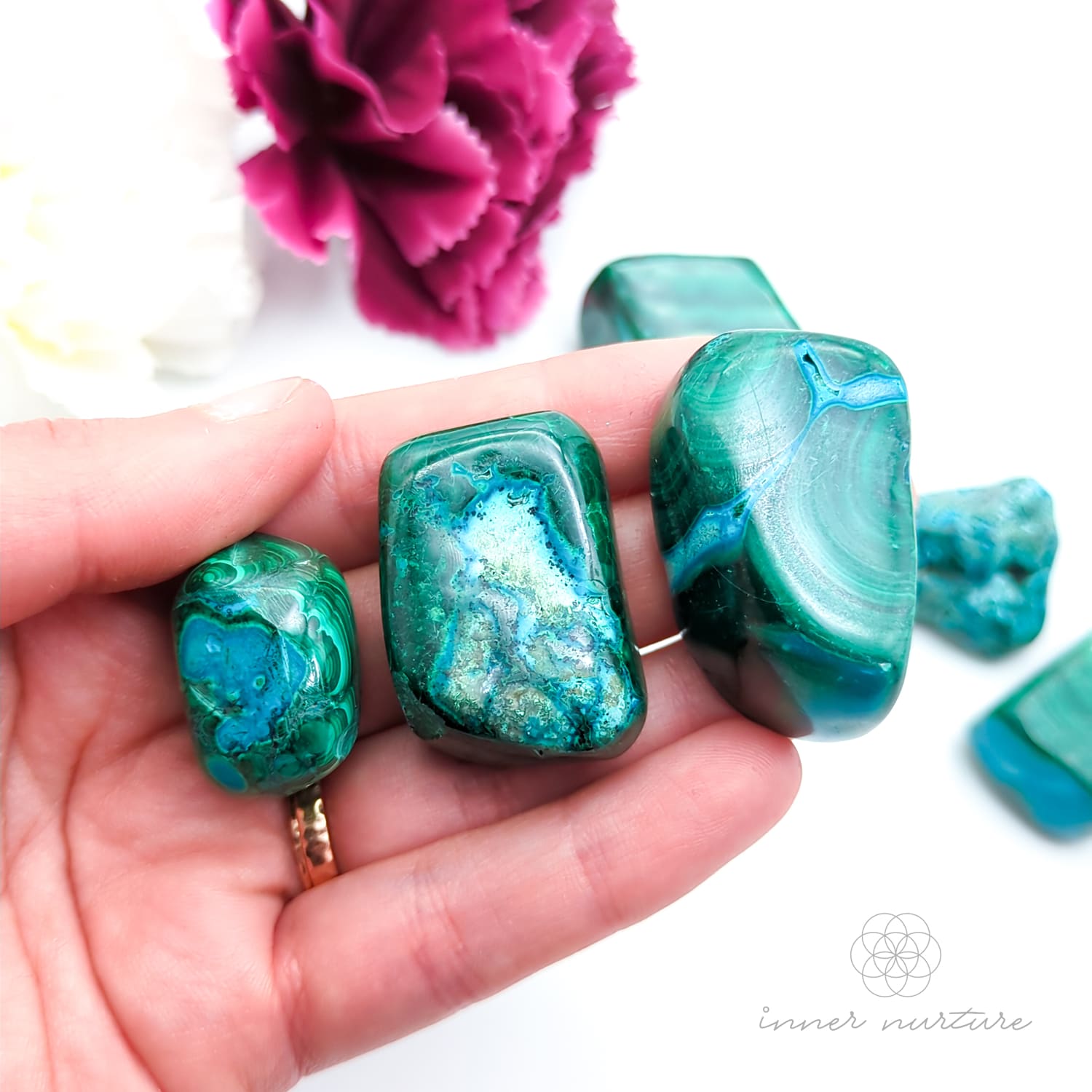 Blue green stones held in hand, with more stones in the background and a white and purple flower. Malacolla sold at Inner Nurture, an online crystal shop in Australia. 