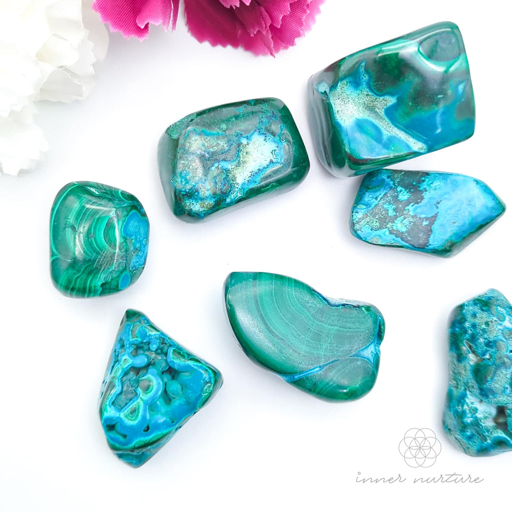 Blue green stone held in hand, with more stones in the background and a white and purple flower. Malacolla sold at Inner Nurture, an online crystal shop in Australia. 