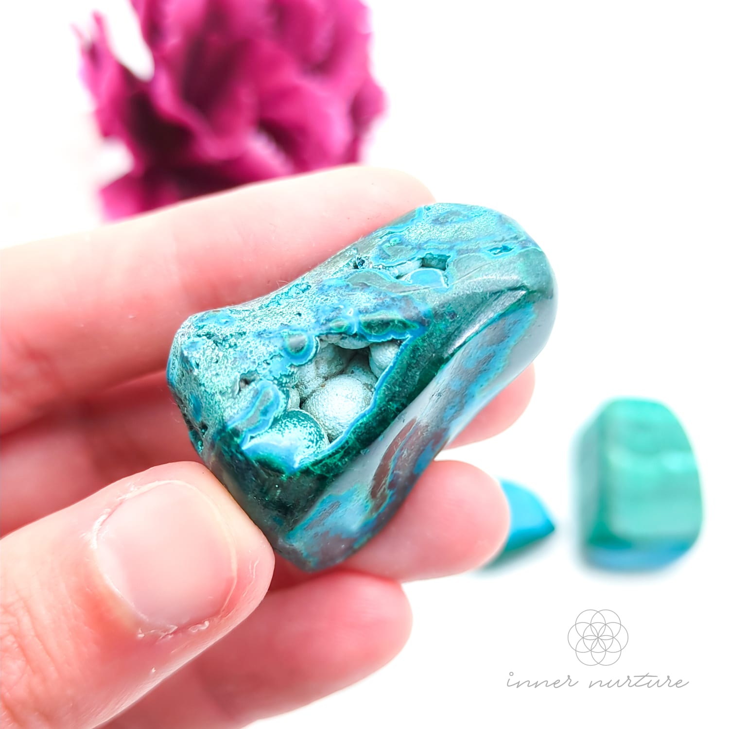 Blue green stone held in hand, with more stones in the background and a white and purple flower. Malacolla sold at Inner Nurture, an online crystal shop in Australia. 