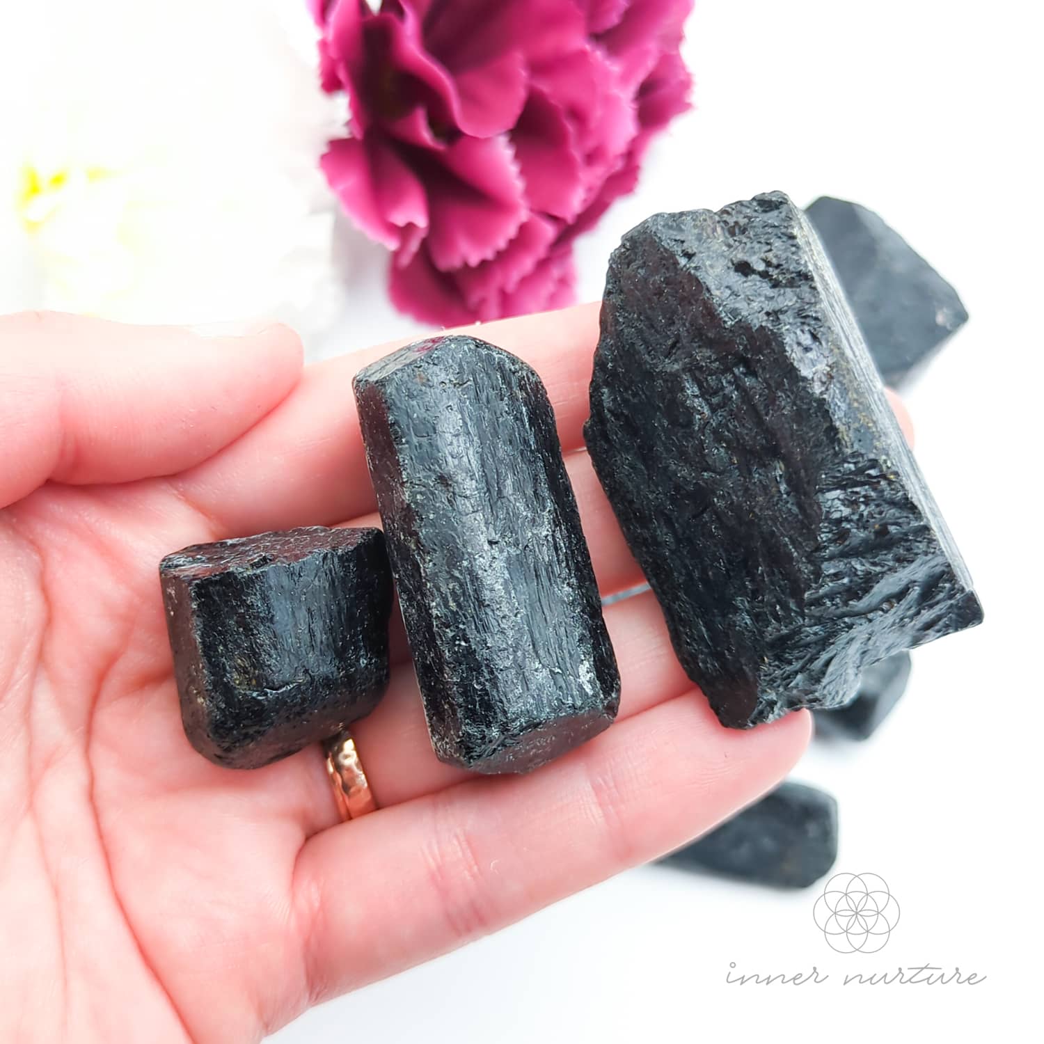 Hand holding a dark crystal with pink flowers in the background. Black Tourmaline available online at Inner Nurture, an Australian crystal store. 