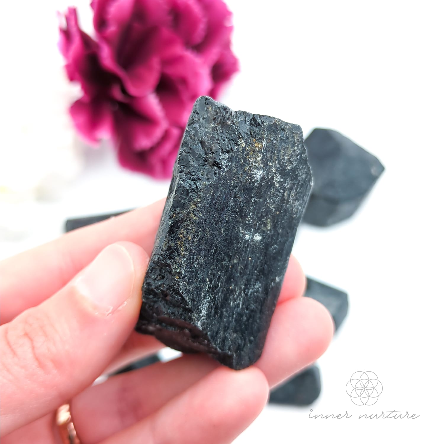 Hand holding a dark crystal with pink flowers in the background. Black Tourmaline available online at Inner Nurture, an Australian crystal store. 