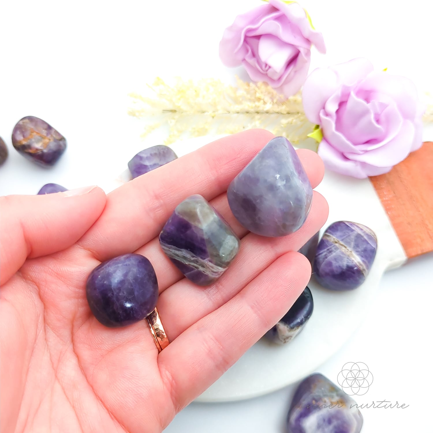Hand holding purple and gray stones with flowers in the background. Amethyst tumbles sold online at Inner Nurture Australia. 