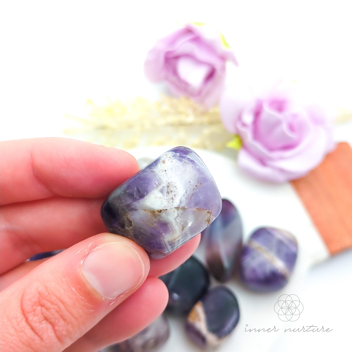 Hand holding purple and gray stones with flowers in the background. Amethyst tumbles sold online at Inner Nurture Australia. 
