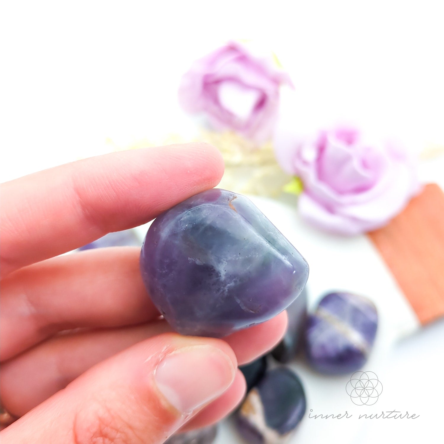 Hand holding purple and gray stones with flowers in the background. Amethyst tumbles sold online at Inner Nurture Australia. 