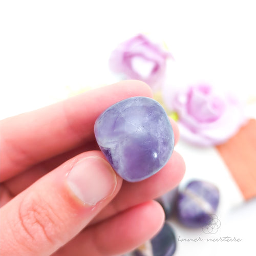 Hand holding purple and gray stones with flowers in the background. Amethyst tumbles sold online at Inner Nurture Australia. 