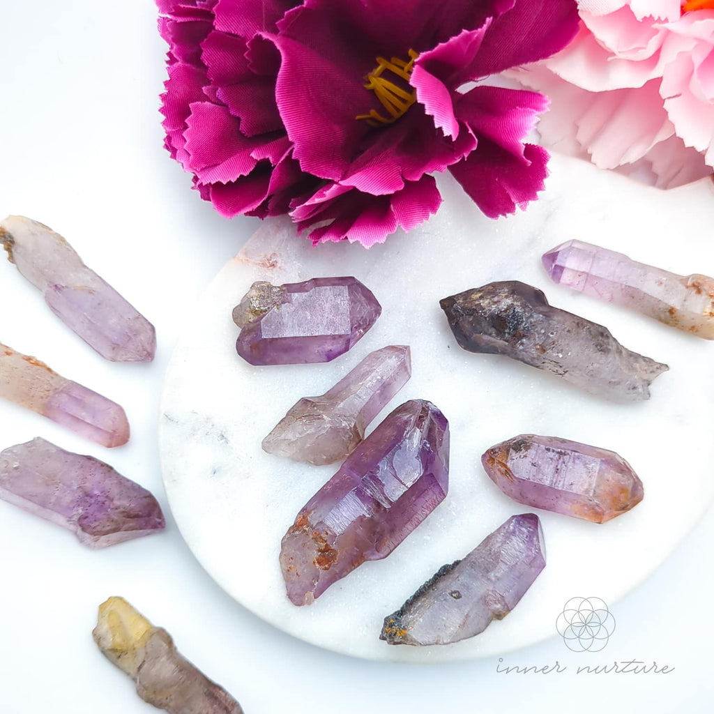 Amethyst crystals on a white surface with pink flowers. Sold online in Australia at Inner Nurture.
