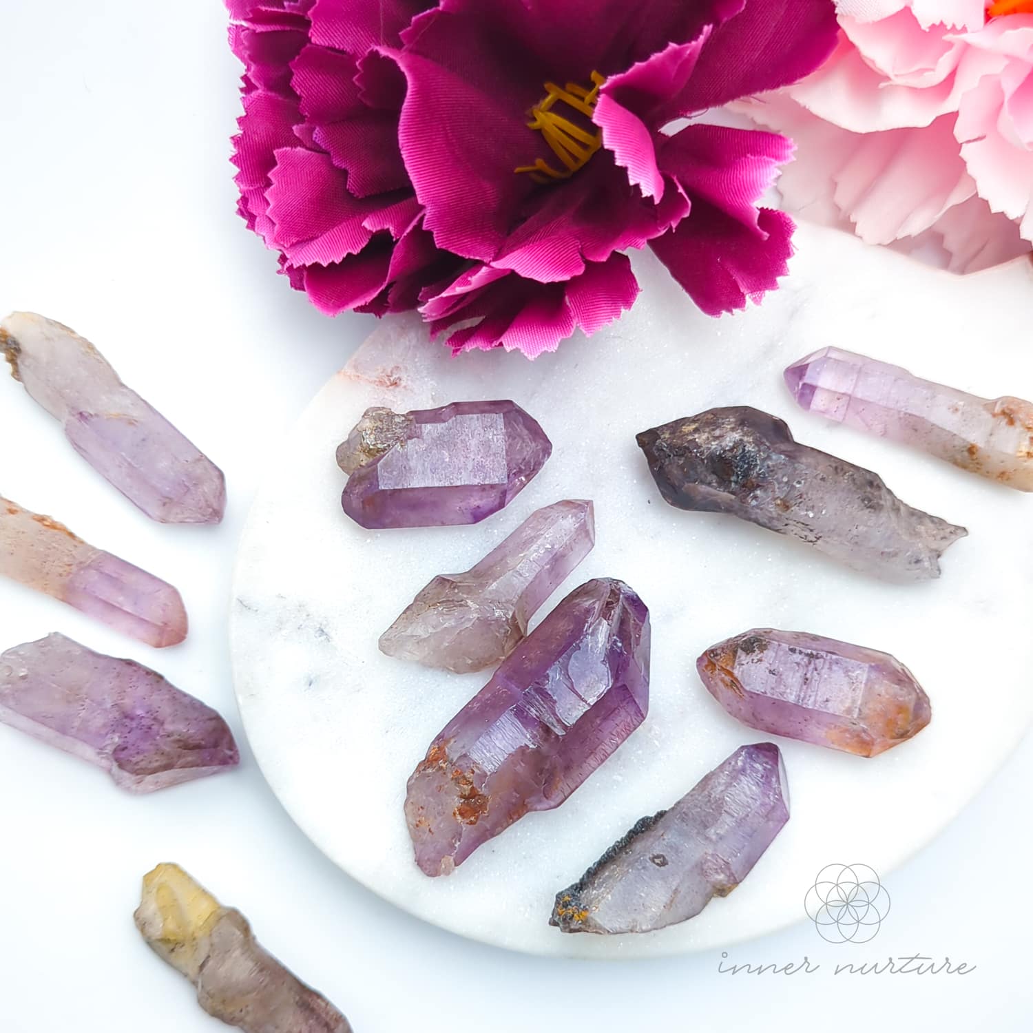 Amethyst crystals on a white surface with pink flowers. Sold online in Australia at Inner Nurture.