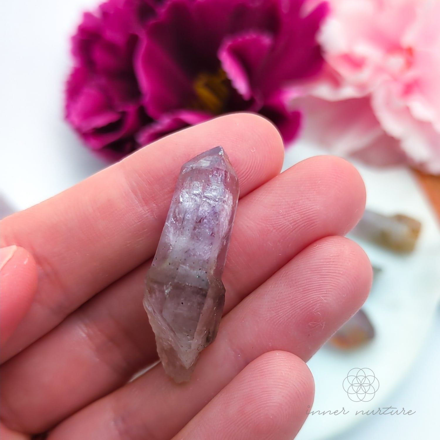 One crystal point held in a hand with pink flowers in the background. Natural Amethyst raw stone sold online in Australia at Inner Nurture. 