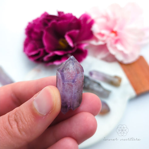 One crystal point held in a hand with pink flowers in the background. Natural Amethyst raw stone sold online in Australia at Inner Nurture. 