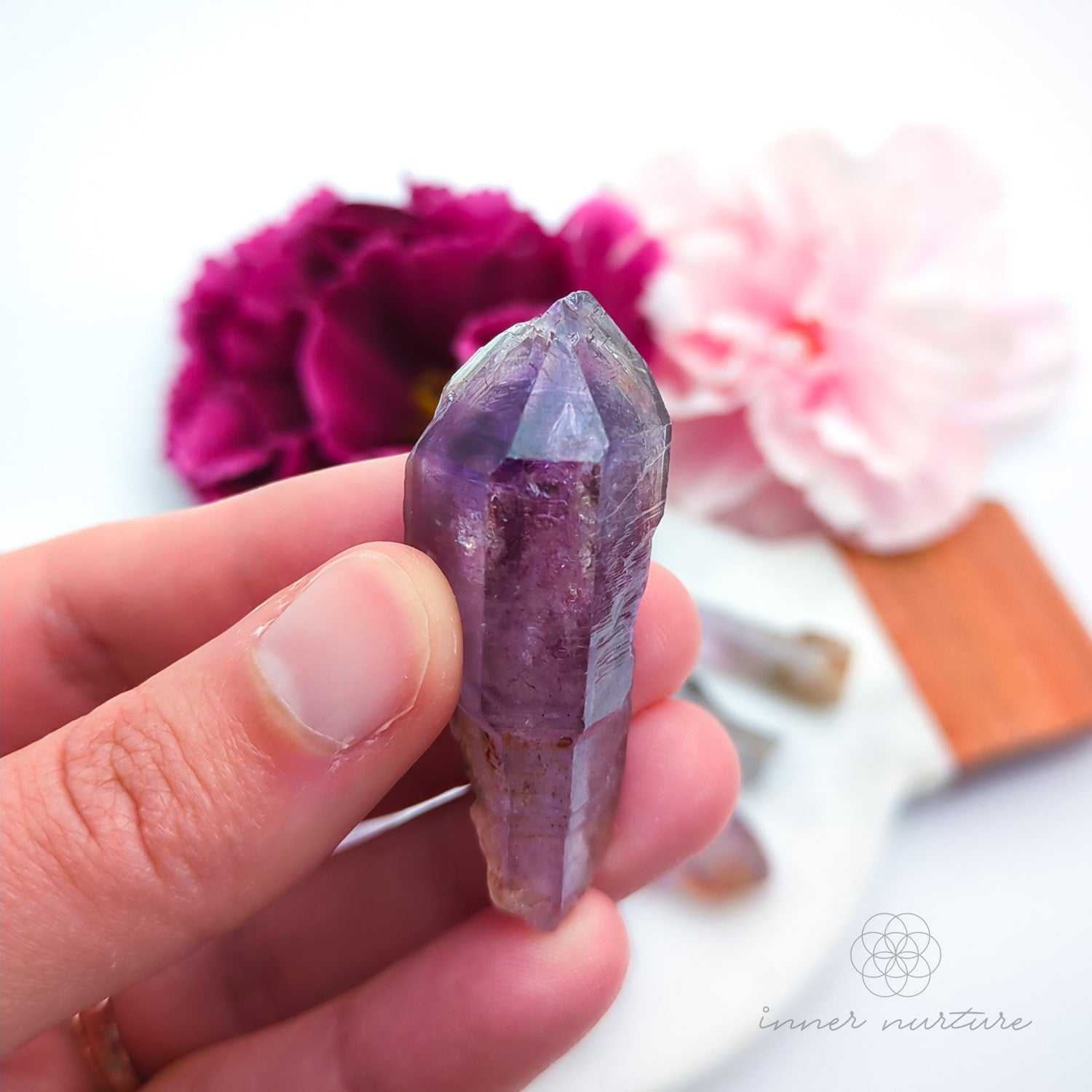 One crystal point held in a hand with pink flowers in the background. Natural Amethyst raw stone sold online in Australia at Inner Nurture. 