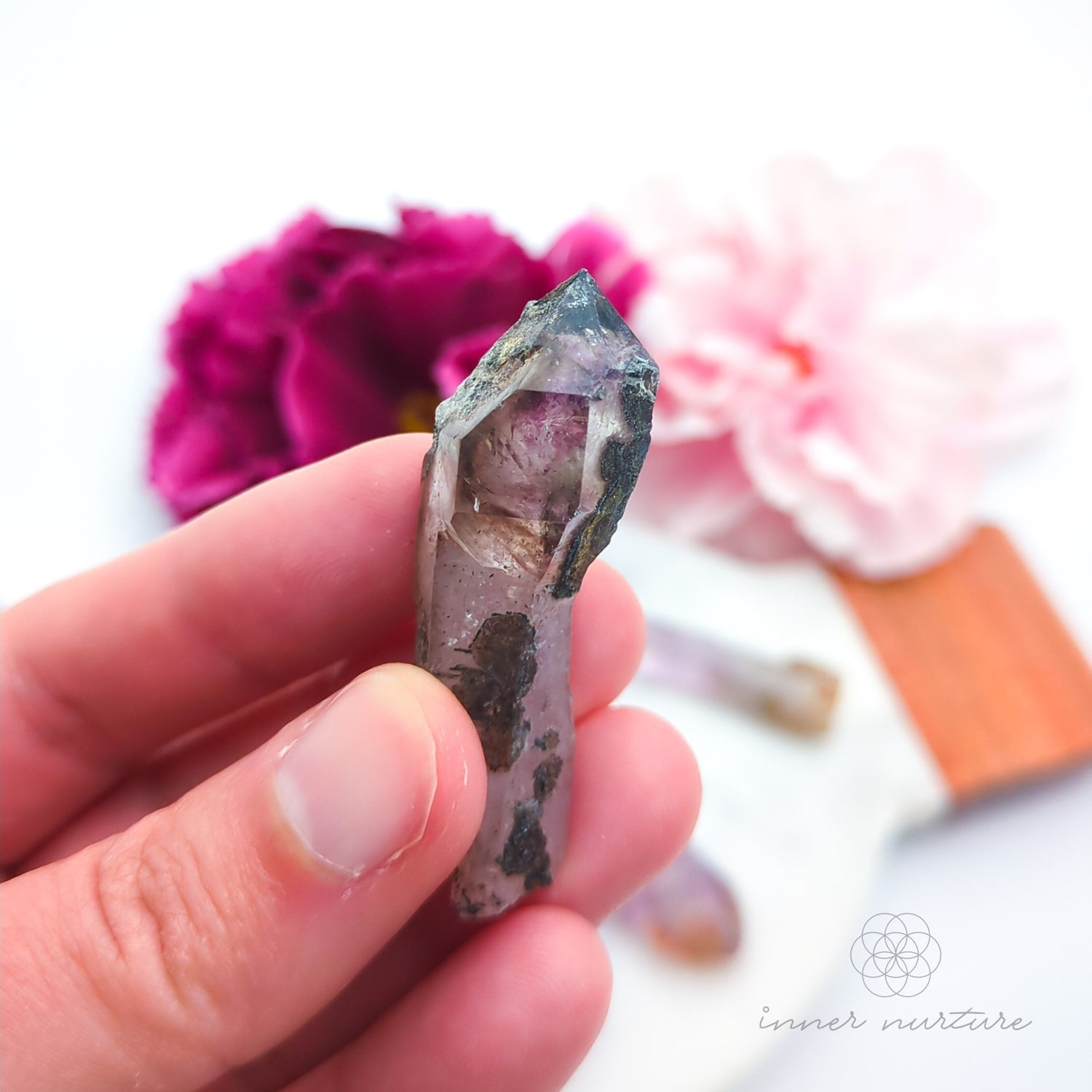 One crystal point held in a hand with pink flowers in the background. Natural Amethyst raw stone sold online in Australia at Inner Nurture. 