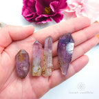 Four crystal points held in a hand with pink flowers in the background. Natural Amethyst raw stone sold online in Australia at Inner Nurture. 