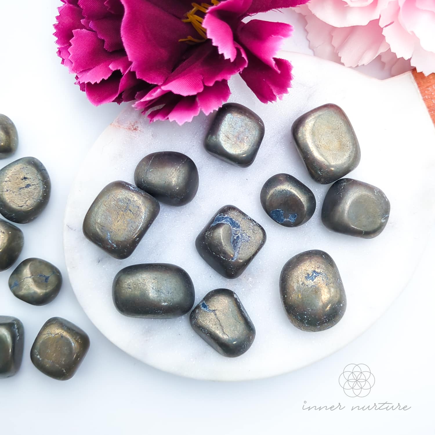 Decorative stones on a white surface with pink flowers in the background. Pyrite tumbles for sale online at Inner Nurture