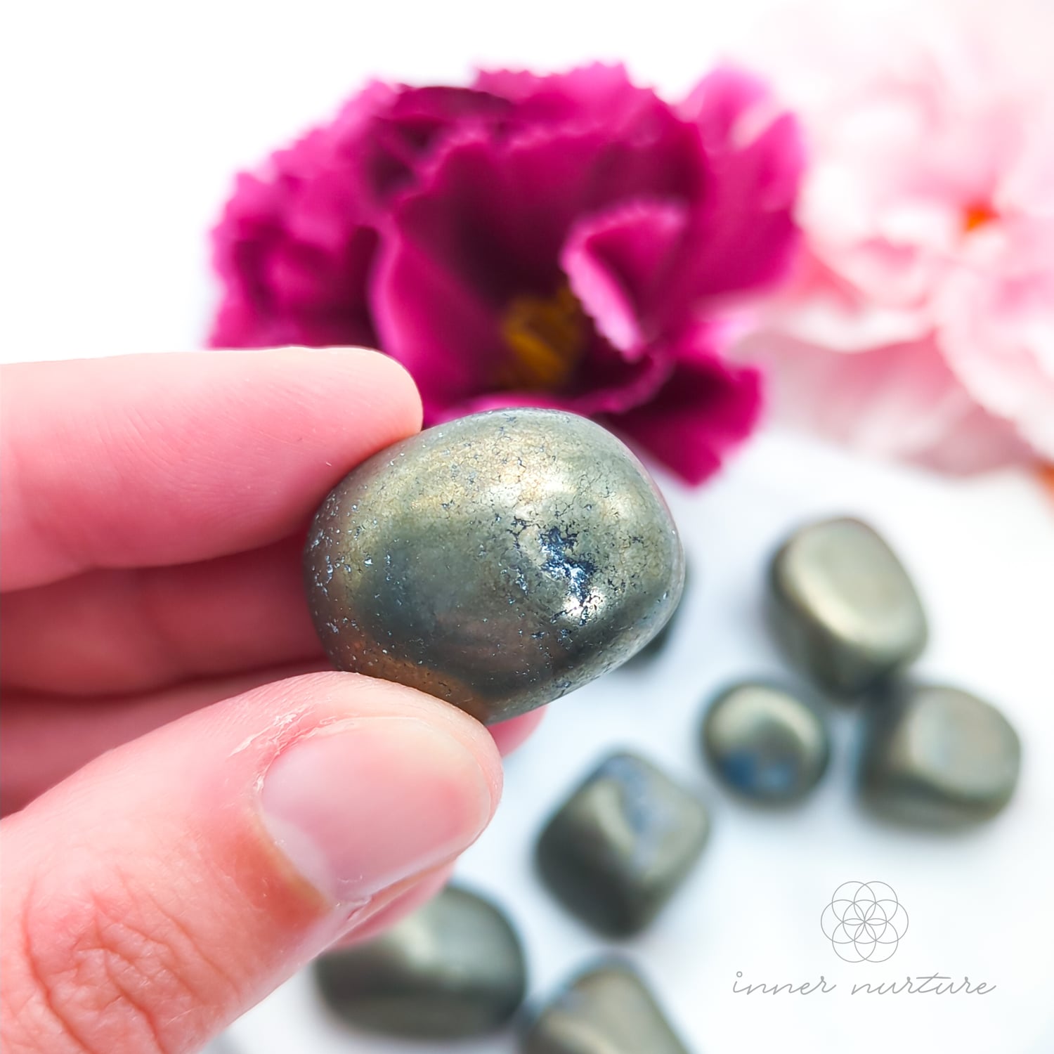Hand holding a metallic golden stone with flowers in the background. Pyrite tumbles for sale online at Inner Nurture