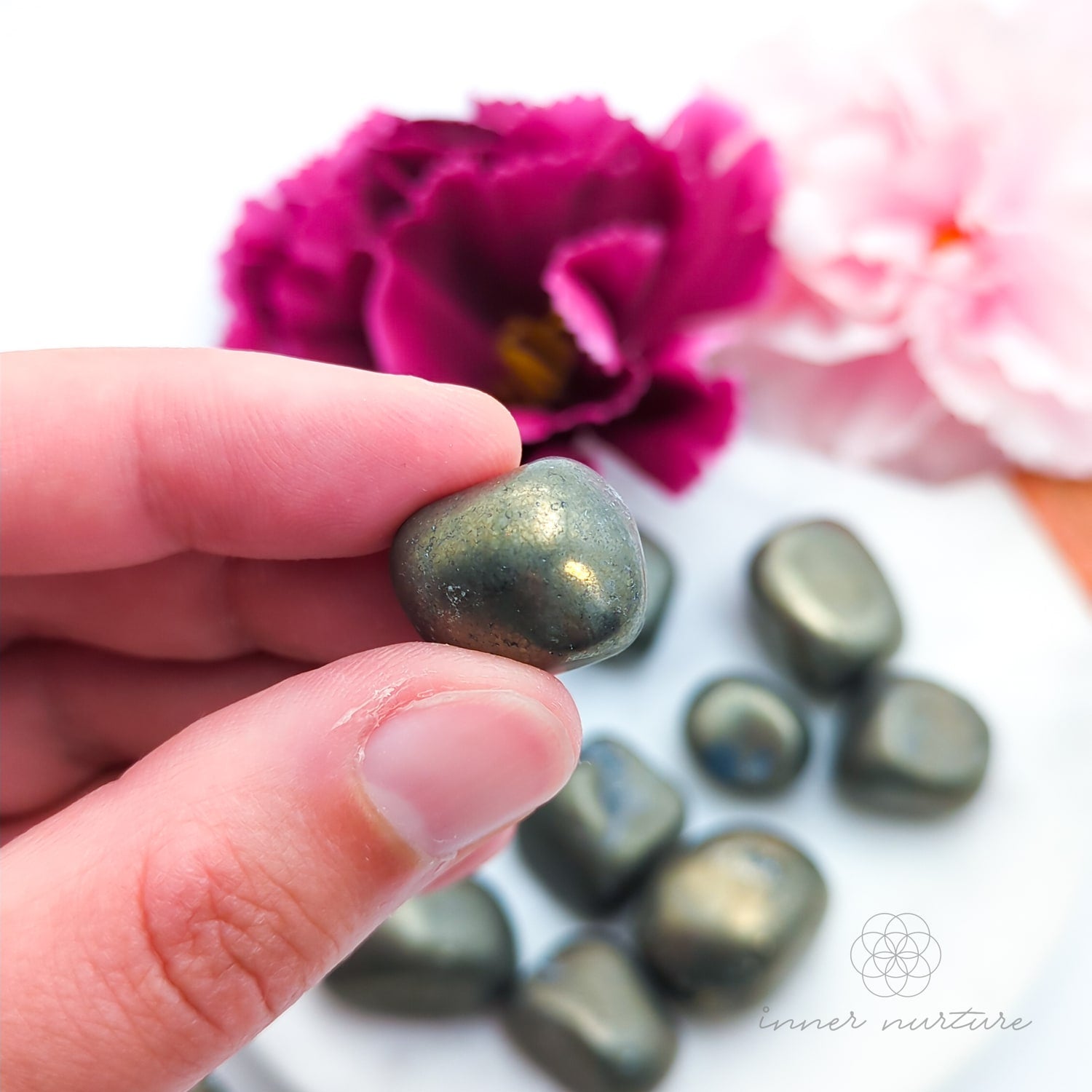 Hand holding a metallic golden stone with flowers in the background. Pyrite tumbles for sale online at Inner Nurture