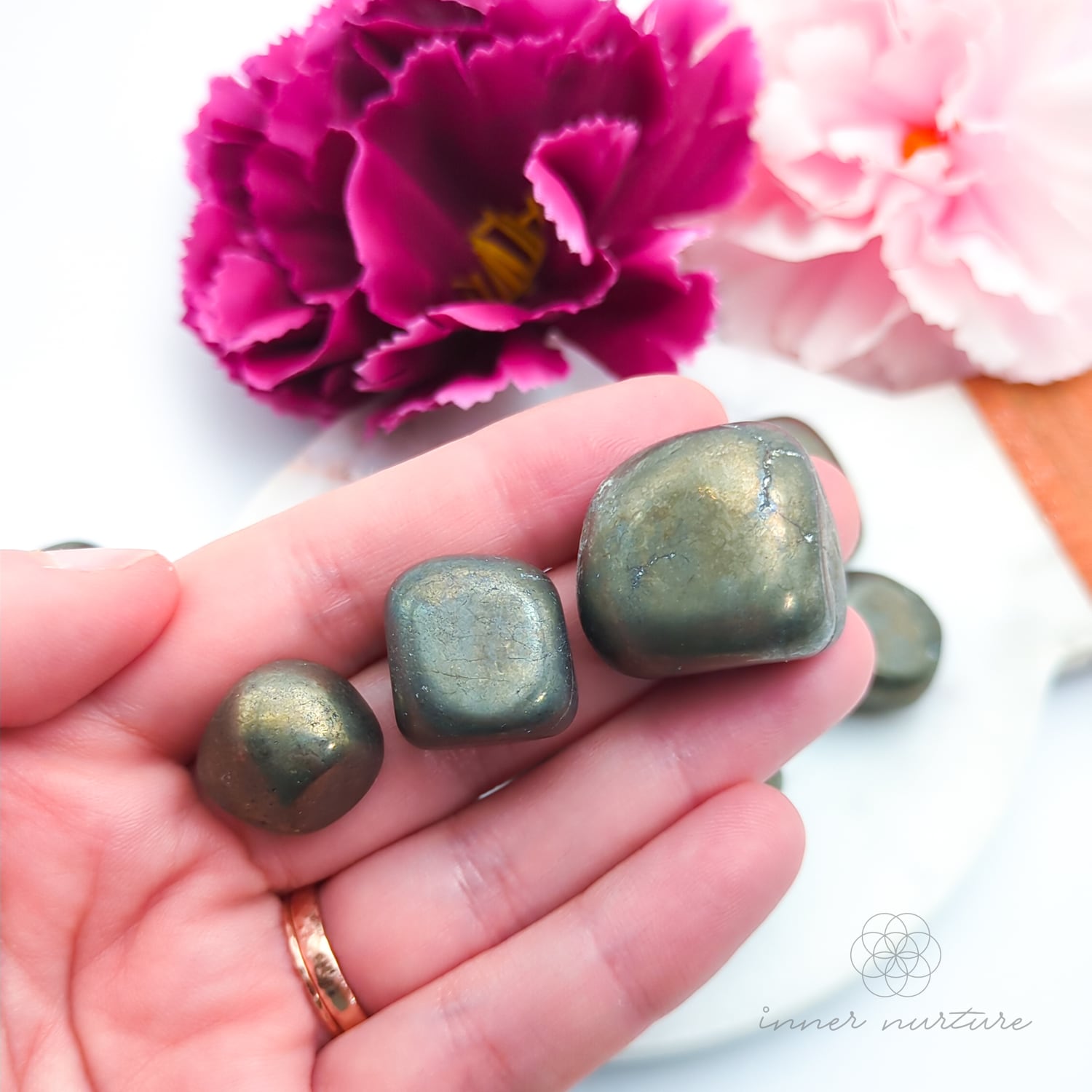 Hand holding a metallic golden stone with flowers in the background. Pyrite tumbles for sale online at Inner Nurture