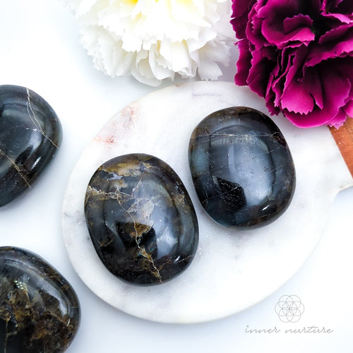 Hand holding a labradorite stone with a pink flower in the background. Labradorite stones available to buy from Inner Nurture, an online crystal shop in australia.