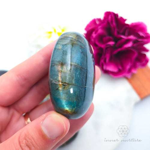 Hand holding a labradorite stone with a pink flower in the background. Labradorite stones available to buy from Inner Nurture, an online crystal shop in australia.