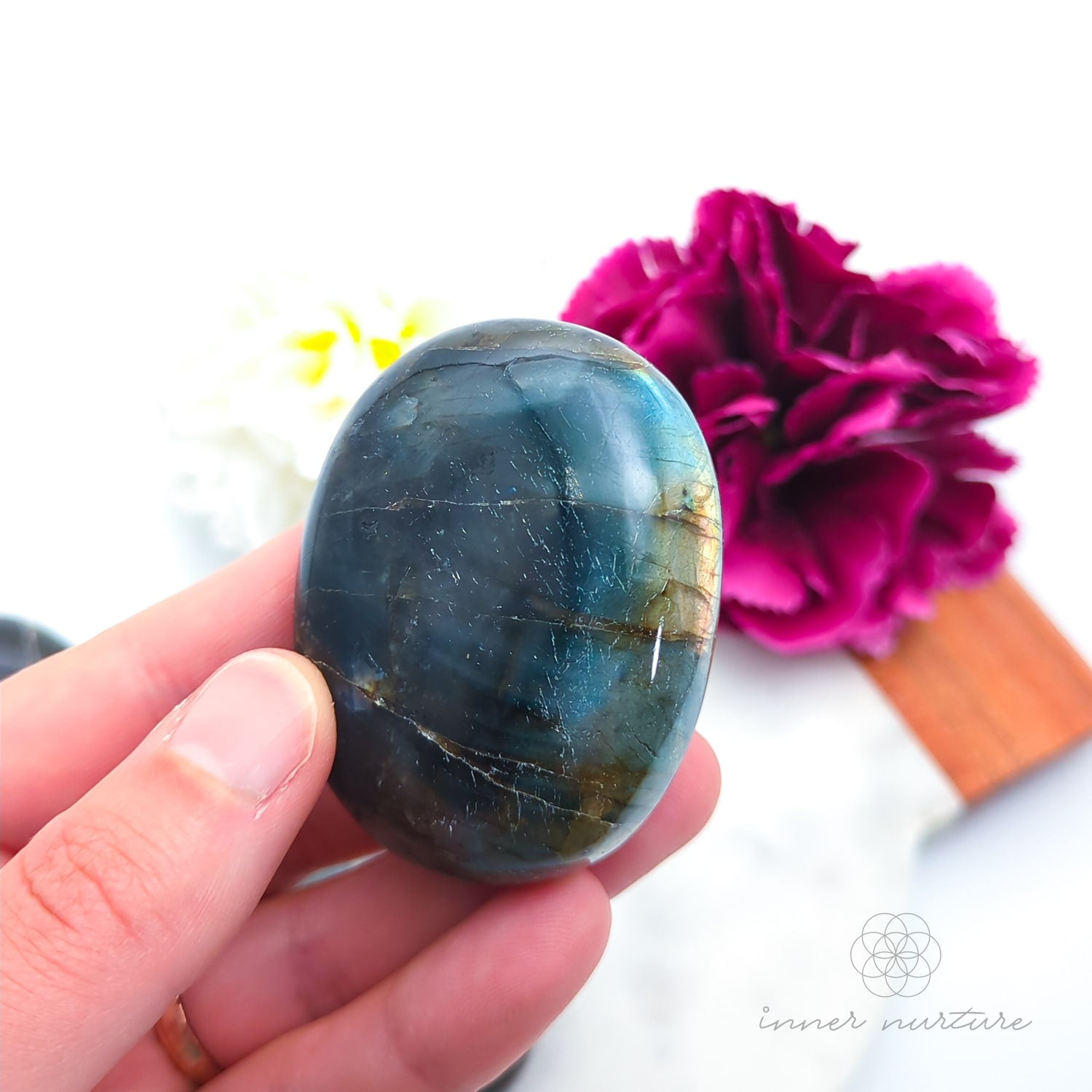 Hand holding a labradorite stone with a pink flower in the background. Labradorite stones available to buy from Inner Nurture, an online crystal shop in australia.