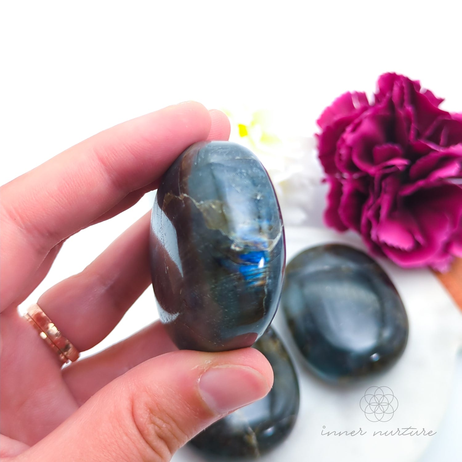 Hand holding a labradorite stone with a pink flower in the background. Labradorite stones available to buy from Inner Nurture, an online crystal shop in australia.