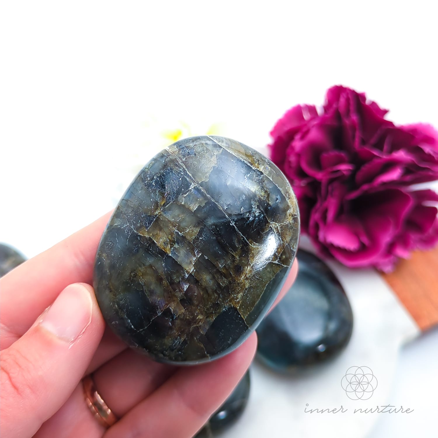 Hand holding a labradorite stone with a pink flower in the background. Labradorite stones available to buy from Inner Nurture, an online crystal shop in australia.