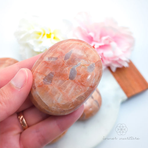 Hand holding a pink stone with floral background. Pink/Peach moonstone palm stone available at Inner Nurture, an online crystal shop in Australia. 