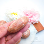 Hand holding a pink stone with floral background. Pink/Peach moonstone palm stone available at Inner Nurture, an online crystal shop in Australia. 