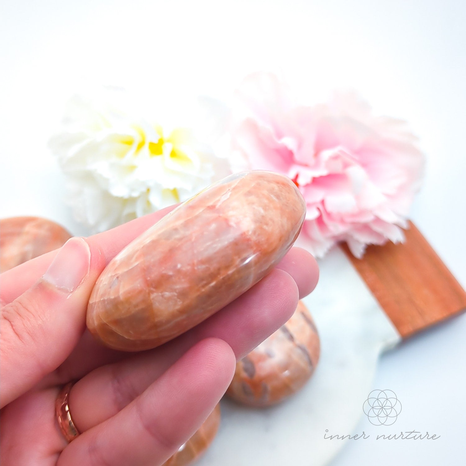 Hand holding a pink stone with floral background. Pink/Peach moonstone palm stone available at Inner Nurture, an online crystal shop in Australia. 