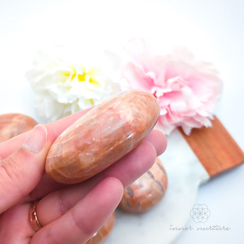 Hand holding a pink stone with floral background. Pink/Peach moonstone palm stone available at Inner Nurture, an online crystal shop in Australia. 